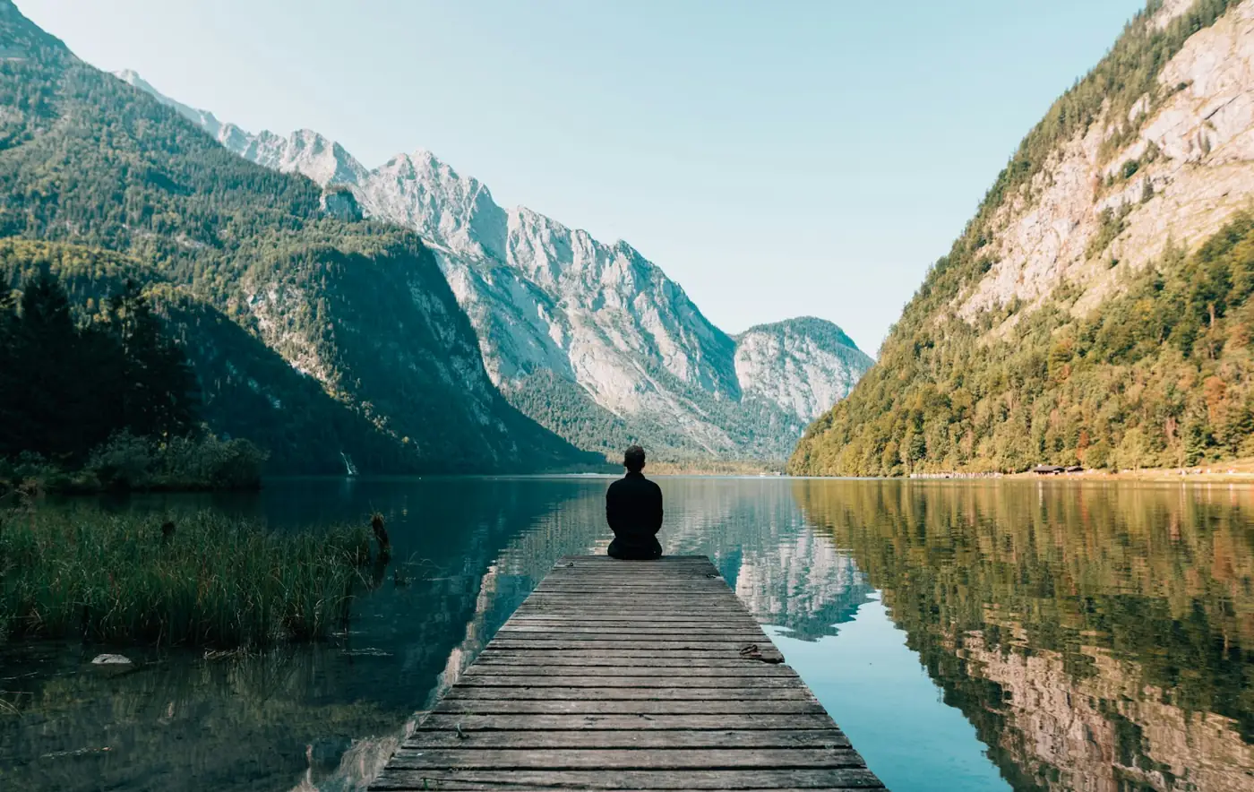 A person sitting at the end of a wooden jetty on a still mountain lake